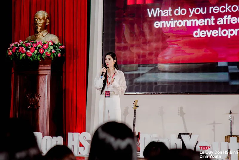 Diễn giả mặc trang phục trắng phát biểu trên sân khấu TEDx, phía sau là màn hình trình chiếu